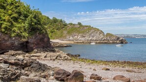A rocky shoreline with a clear view of the sea, a small boat, and distant buildings. - 3 Dolphin Court (Brixham)