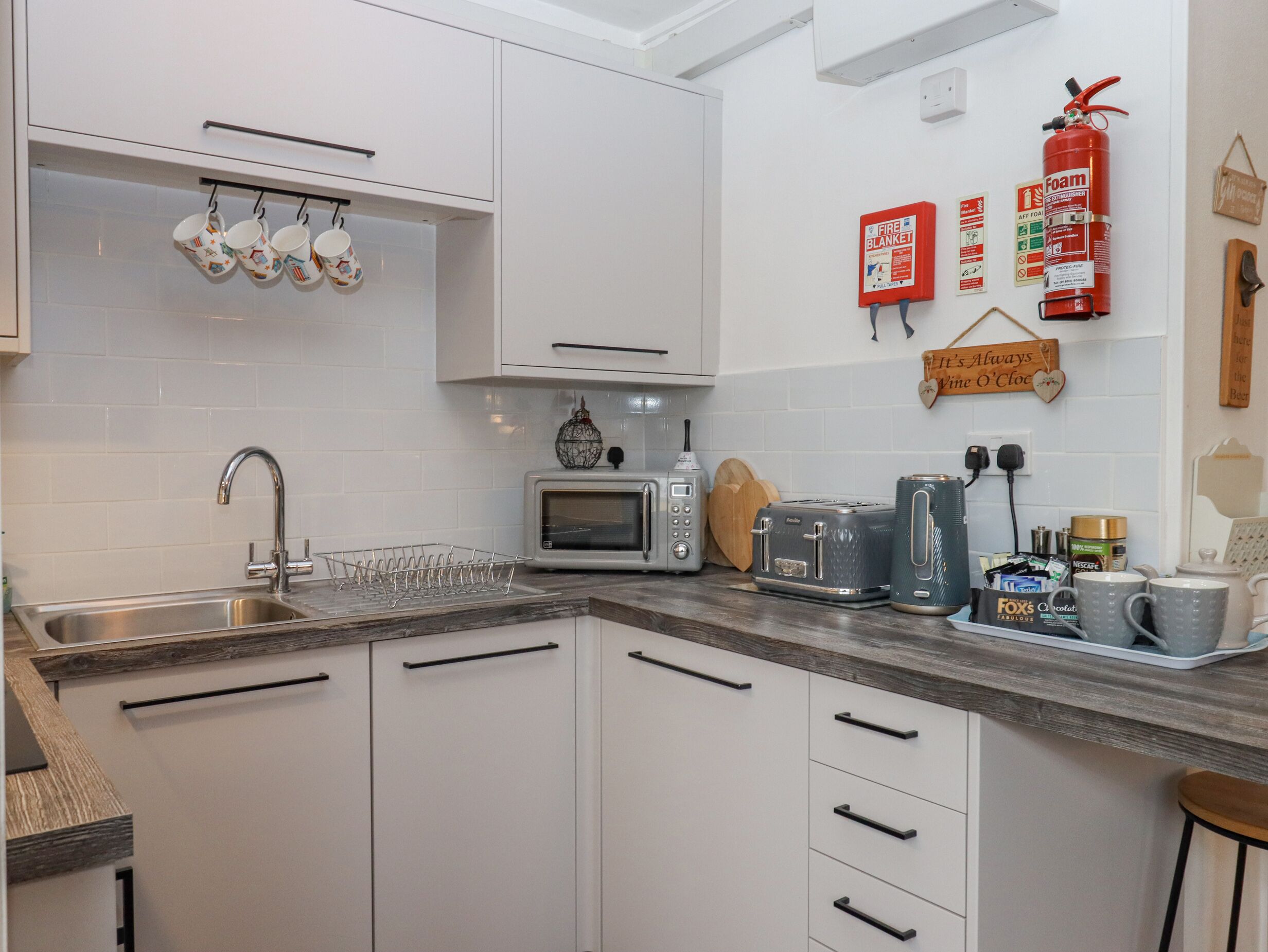 A modern kitchen with white cabinets, a stainless steel sink, a microwave, a toaster, and a coffee machine. There are also fire safety signs on the wall and a hanging mug rack.
