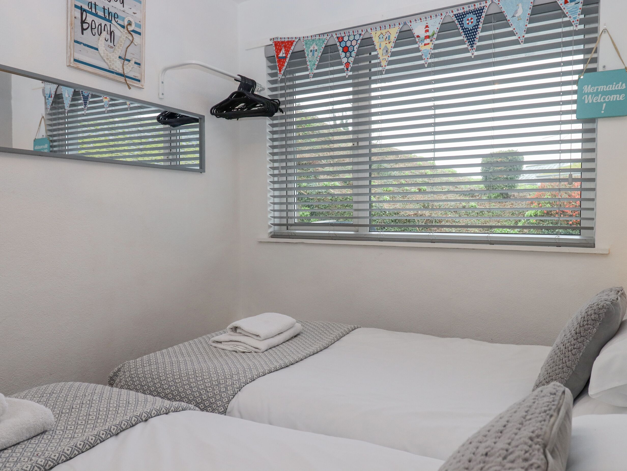 Ein Hotelzimmer mit zwei Betten, einem Fenster mit Jalousien, einem Spiegel und einem hängenden Handtuchhalter.