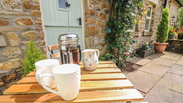 A wooden table with two white mugs and a white pitcher, set against a stone wall with a door and potted plants. - Spens Farm Cottage (Lancaster)