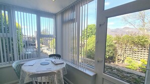 En matplats med ett bord och stolar, stora fönster med persienner och utsikt över en trädgård och berg.