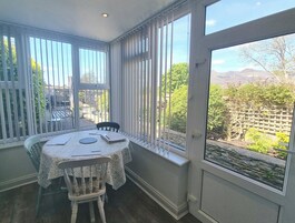 A dining area with a table and chairs, large windows with blinds, and a view of a garden and mountains.