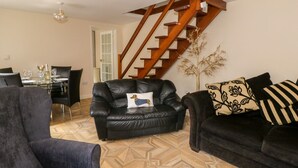 A living room with a black leather sofa, a dining table set with glasses and a bottle, a staircase, and a decorative plant. - Rose Cottage (Lydney)