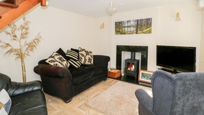 A living room with a black sofa, a grey armchair, a fireplace, a television, and a wooden beam structure. - Rose Cottage (Lydney)
