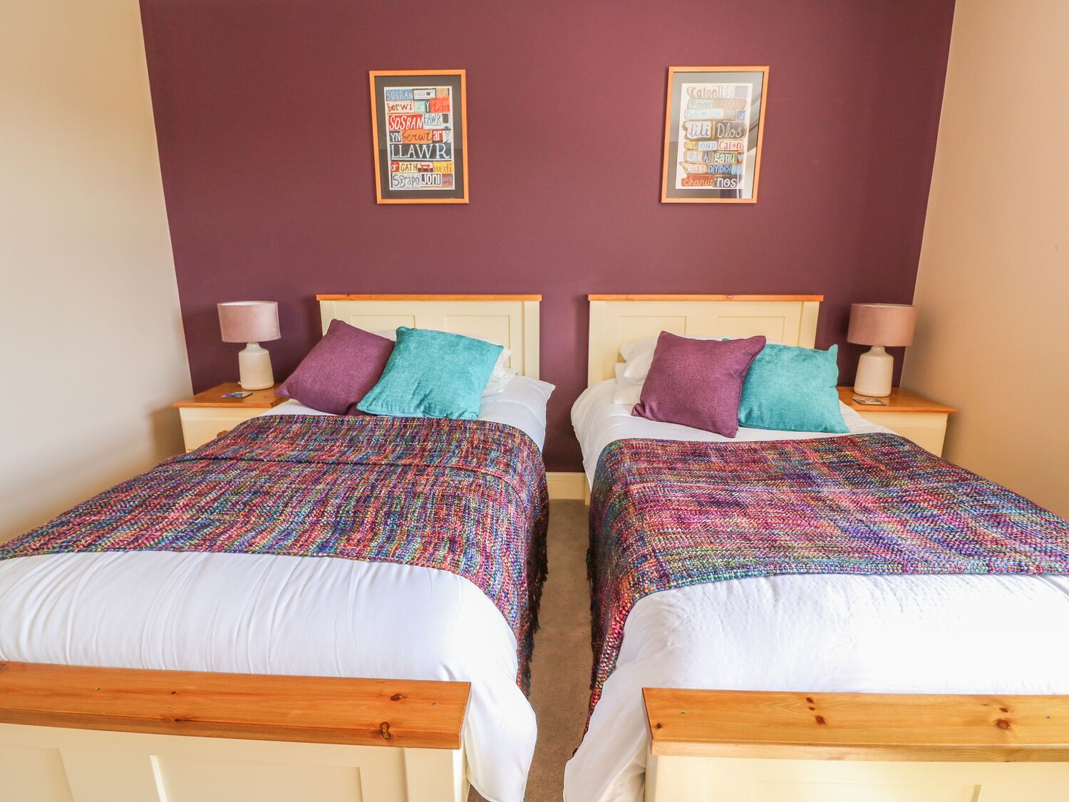 A double room with two beds, each with a wooden headboard, white bedding, and colorful throw pillows. There are bedside tables with lamps and framed posters on the wall above the beds.