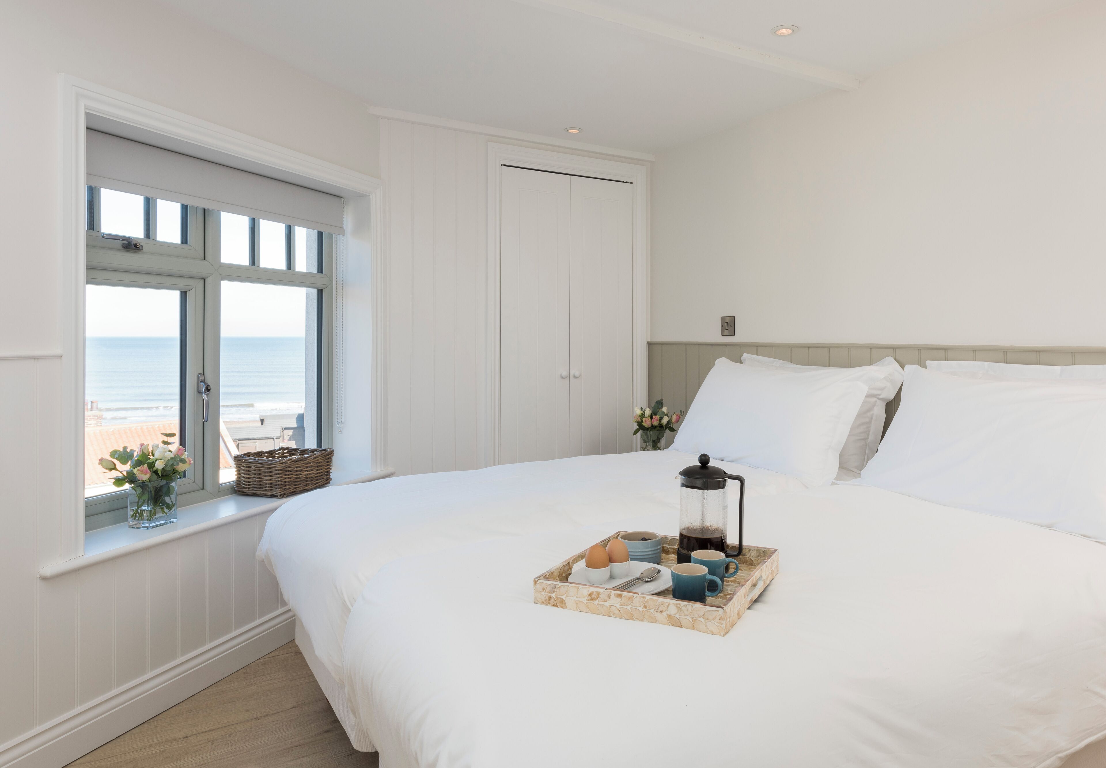 Ein ordentlich bezogenes Bett, ein Tablett mit Frühstücksgegenständen, ein Fenster mit Meerblick und eine geschlossene Tür.