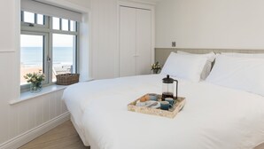 En snyggt bäddad säng med ett brickbord fullt av frukosttillbehör, ett fönster med utsikt över havet och en stängd dörr.
