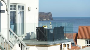 En balkong med ett glasräcke som vetter mot havet, ett bord med en flaska och frukt, och en skylt med texten ”Utsikt över stranden”.