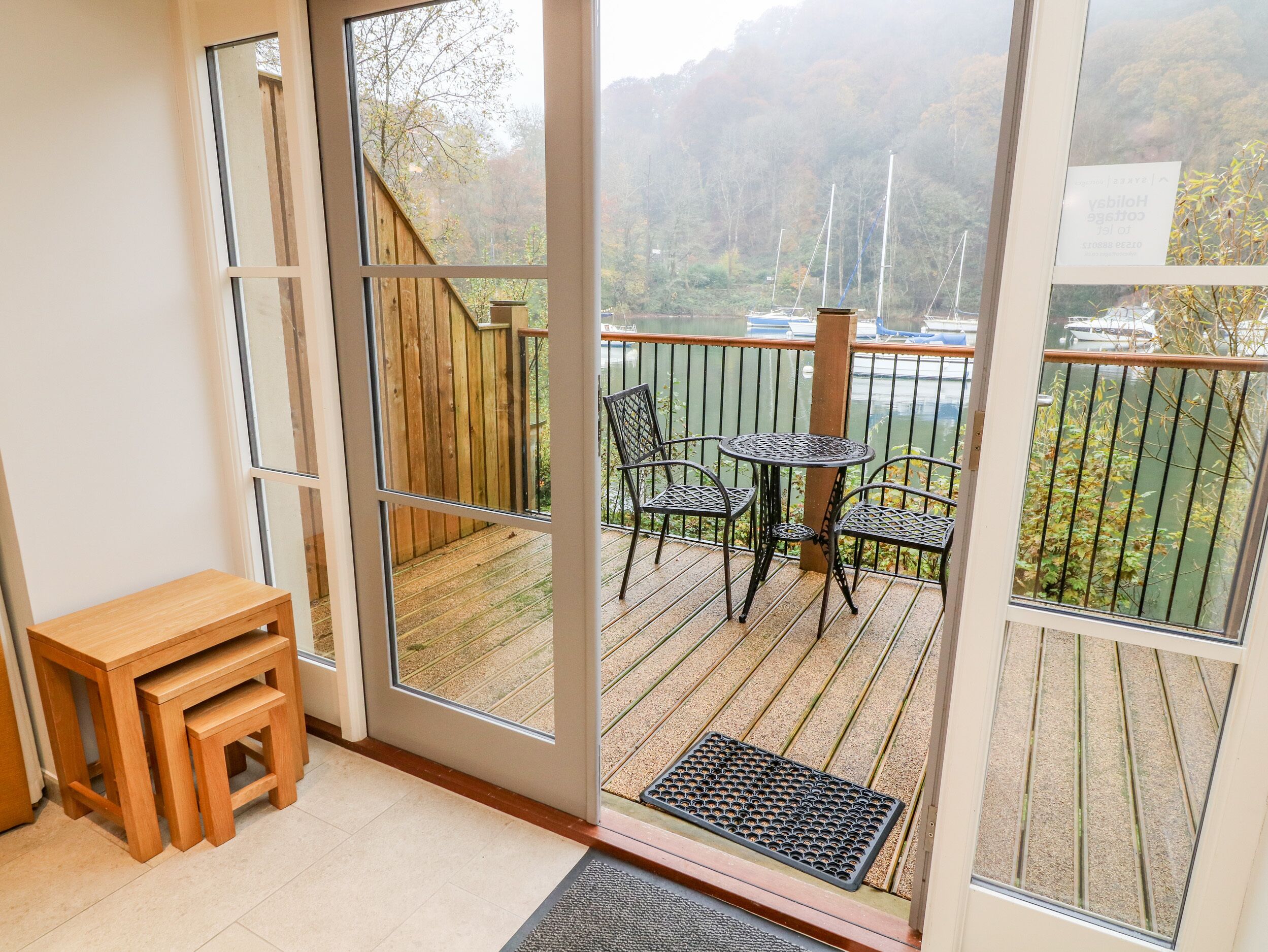 A room with a sliding glass door leading to a balcony with a table and chairs, a wooden deck, and a view of boats on water.