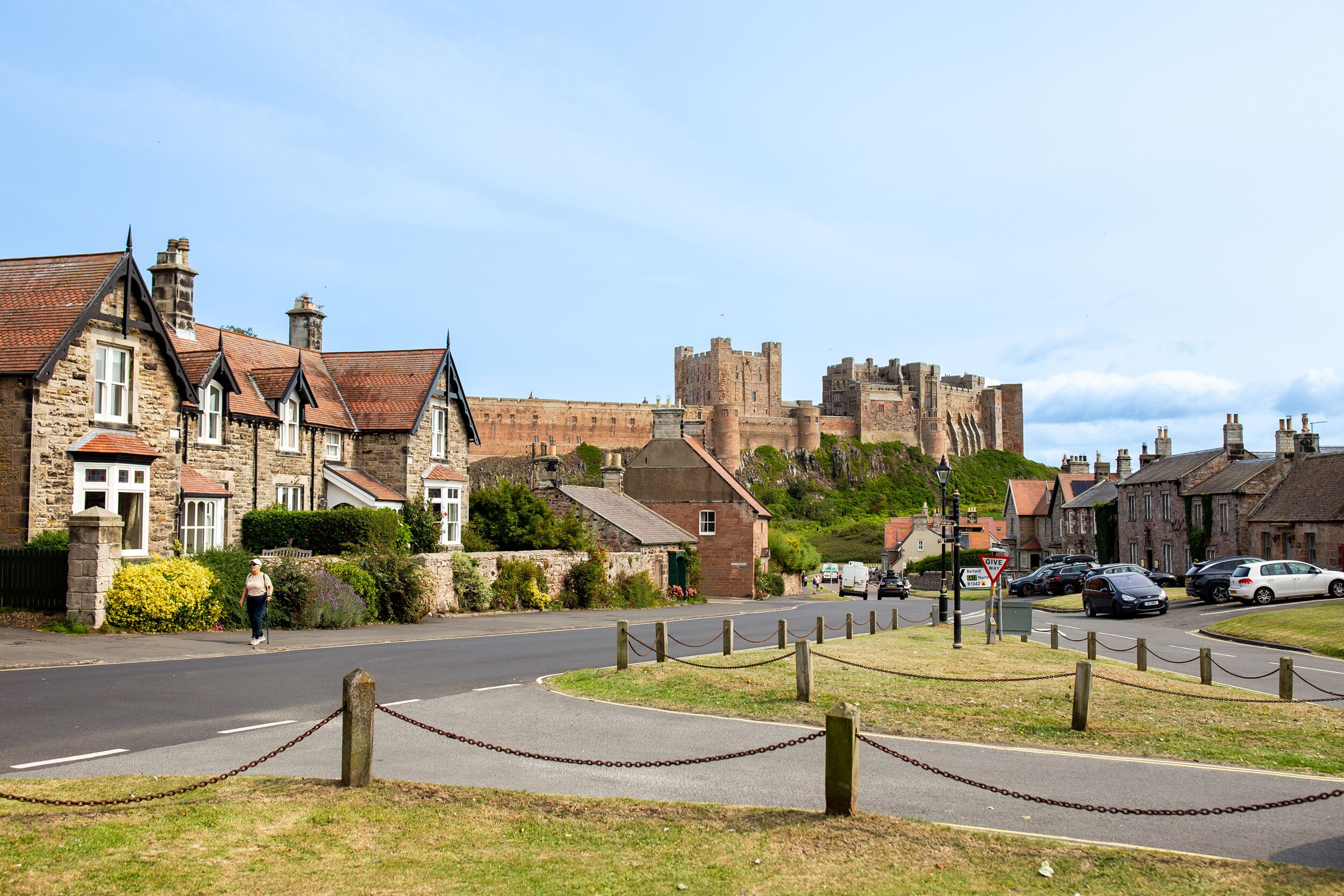 A historic stone building with a traditional roof, a paved road with a chain barrier, and a castle in the background.