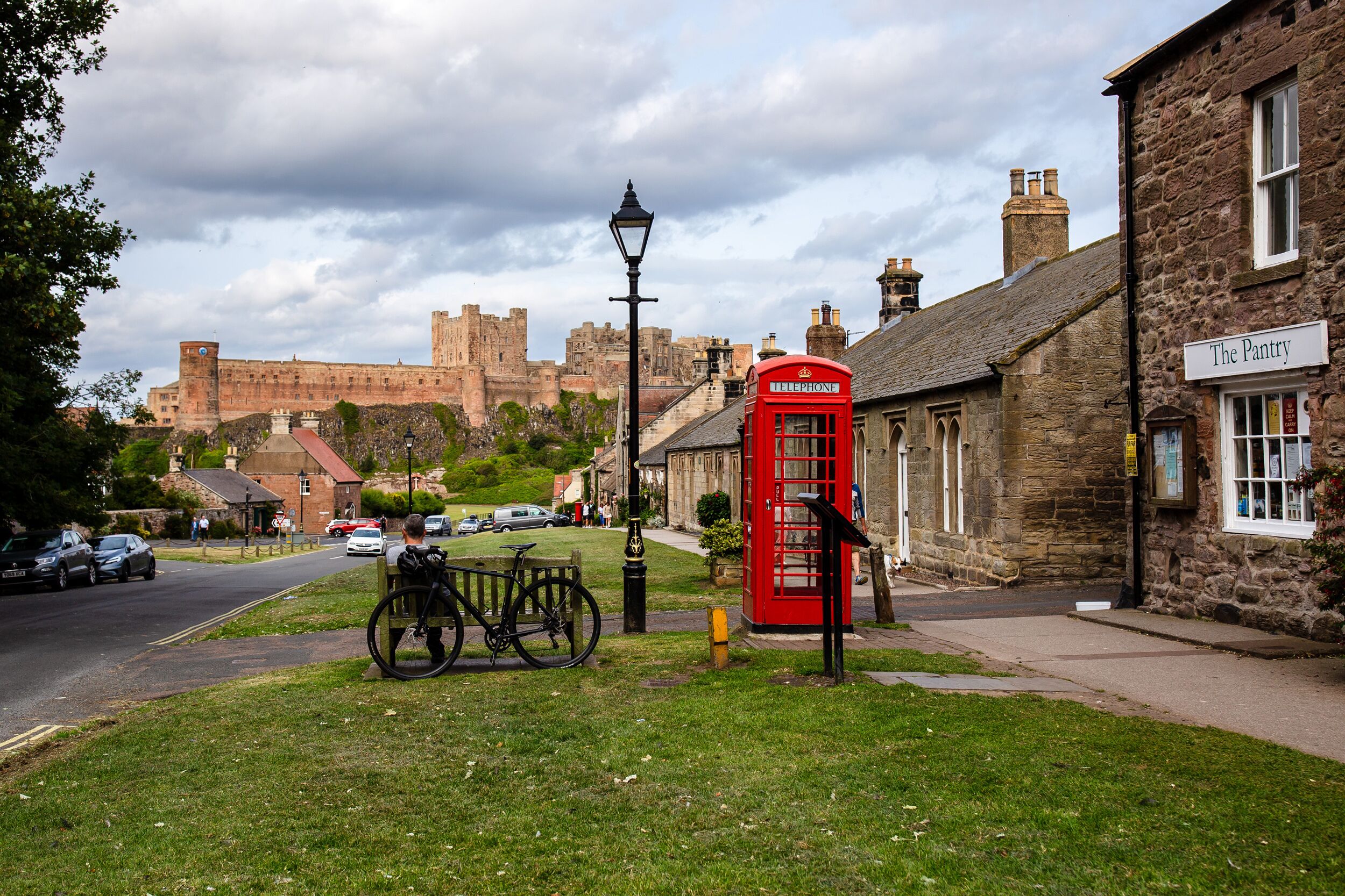 A traditional British street with a red telephone booth, a stone building labeled 'The Pantry,' and a historic castle in the background.