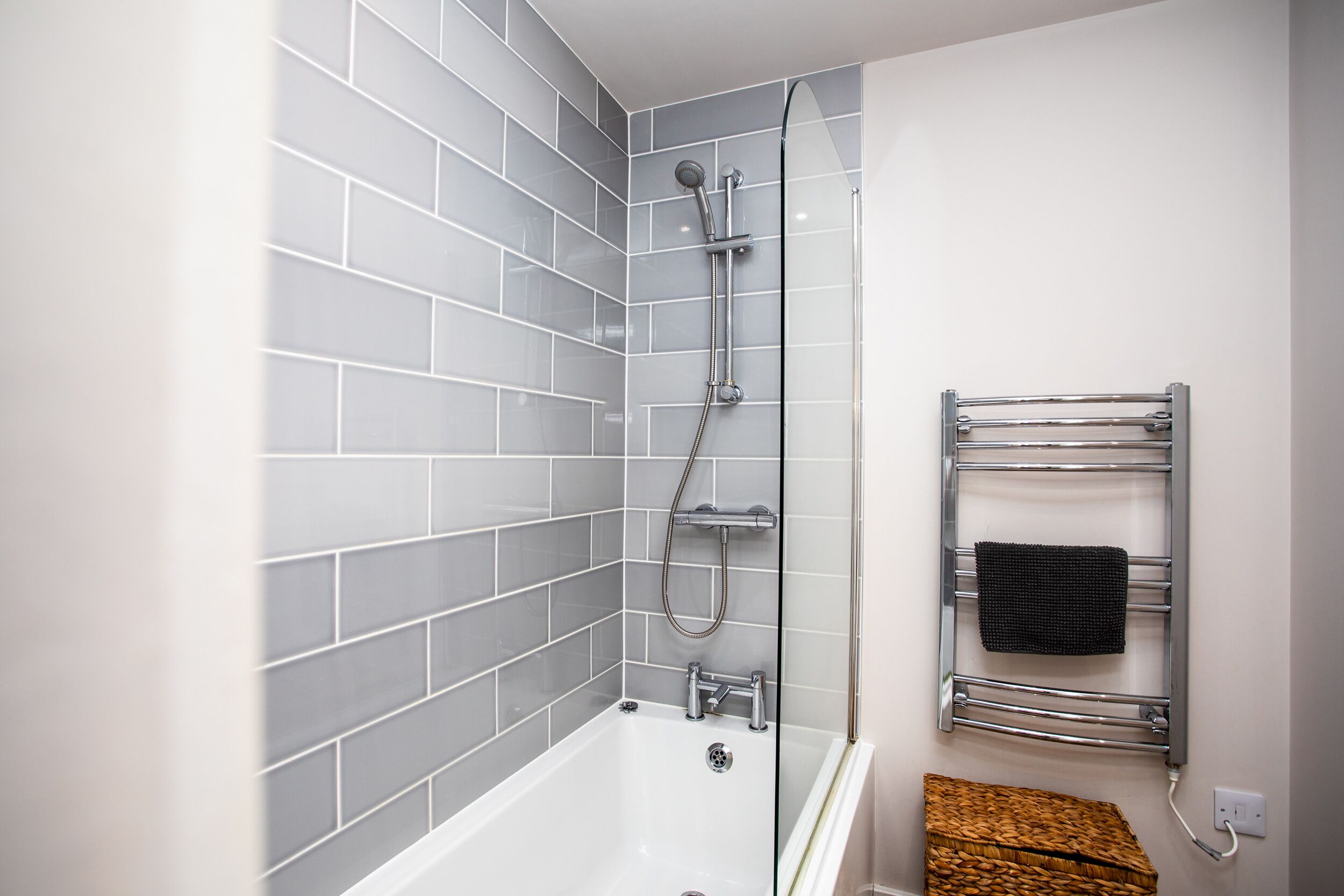 A modern bathroom with grey tiled walls, a white bathtub, a glass-enclosed shower, and a towel rack with a black towel.