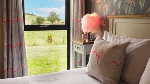 Un dormitorio con un ventanal grande, una mesita de noche con una lámpara de plumas rosadas y una pared con estampado floral.