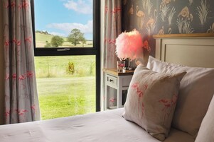 Un dormitorio con un ventanal grande, una mesita de noche con una lámpara de plumas rosadas y una pared con estampado floral.