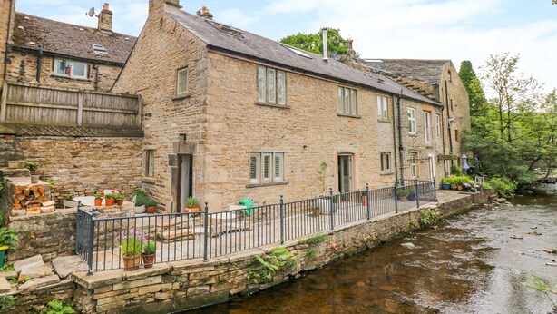 A stone building with a river in front, a wooden fence, and potted plants. - River Cottage Hayfield (High Peak)