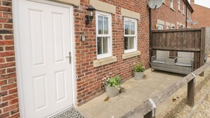 A red brick house with a white door, two windows, and a small outdoor seating area with a wooden fence. - Crossing Cottage (Whitby)