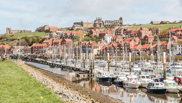 A marina with boats docked, a river, and buildings in the background. - Crossing Cottage (Whitby)