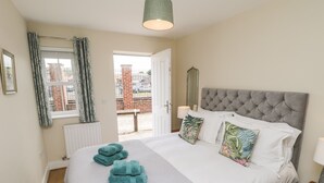 A bedroom with a large bed, a grey upholstered headboard, and a view of the outside through an open door. - Crossing Cottage (Whitby)