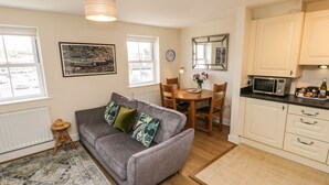 A cozy living room with a grey sofa, a small wooden stool, and a patterned rug. There's a dining area with a wooden table and chairs, and a kitchen with white cabinets and a microwave. - Crossing Cottage (Whitby)