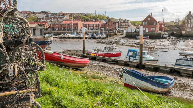 Un pueblo costero con barcos amarrados en un muelle, rodeado de edificios de ladrillo rojo y un primer plano con césped.