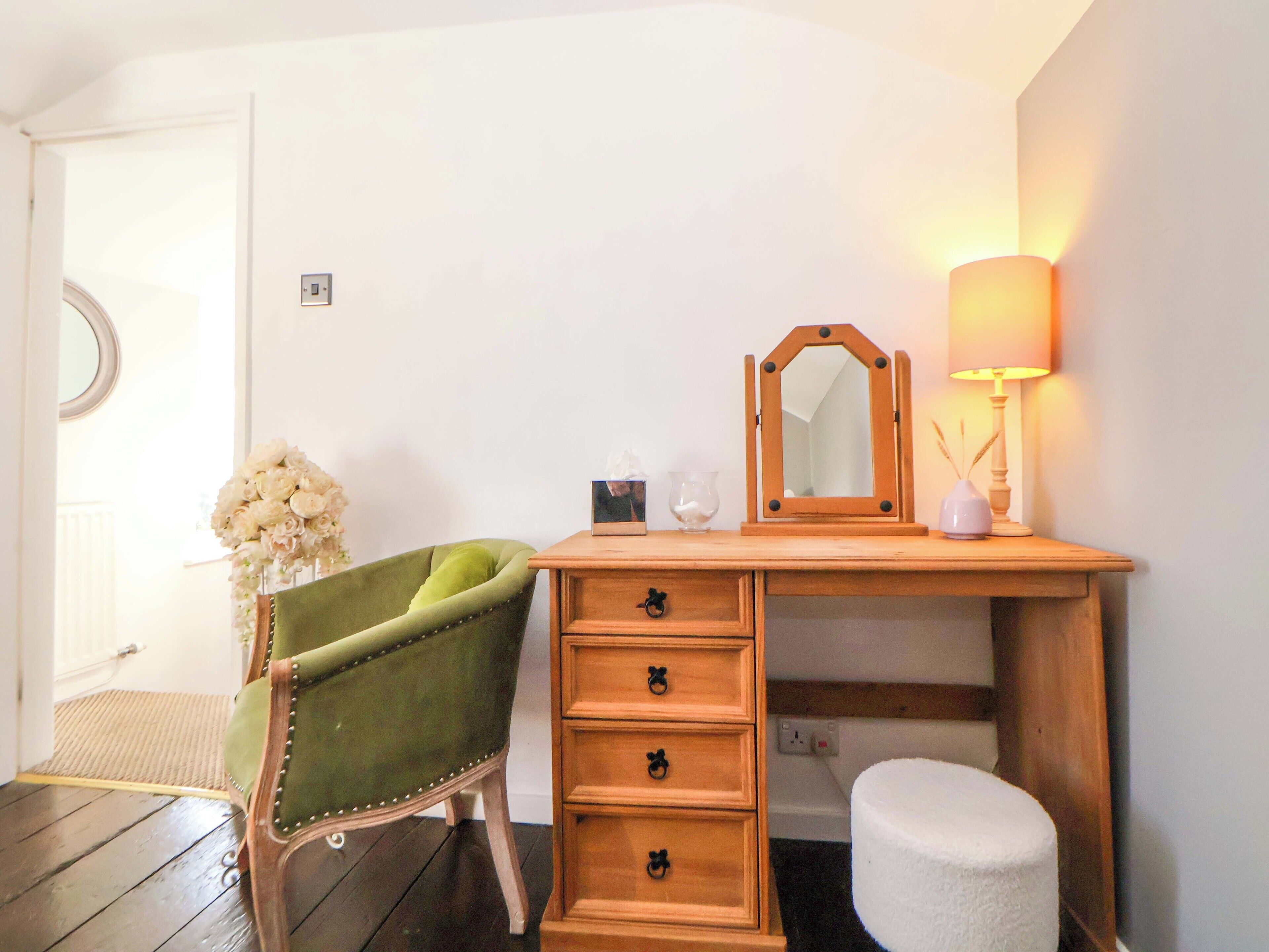 A wooden dresser with a mirror, a lamp, and a green chair in a room with white walls and wooden flooring.