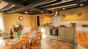 A kitchen with a dining area, wooden chairs, a table set for two, a clock on the wall, and a tiled backsplash.
