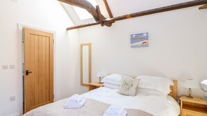 A bedroom with a wooden ceiling, a bed with white linens, a wooden nightstand, a bedside table with a lamp, and a wall-mounted mirror. - Sparrow Barn (Norwich)