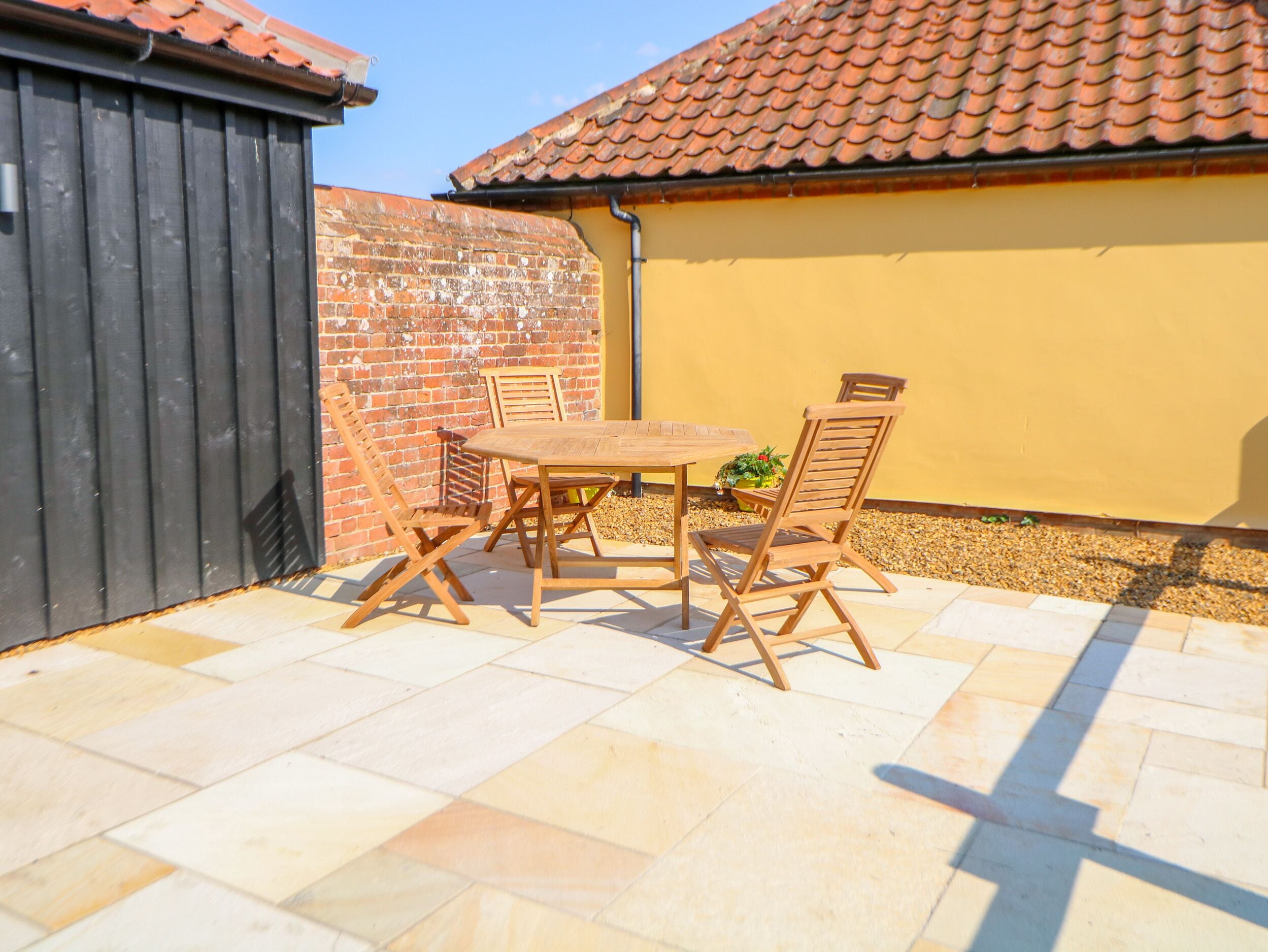 A patio with a wooden table and chairs, a brick wall, and a yellow building with a tiled roof.