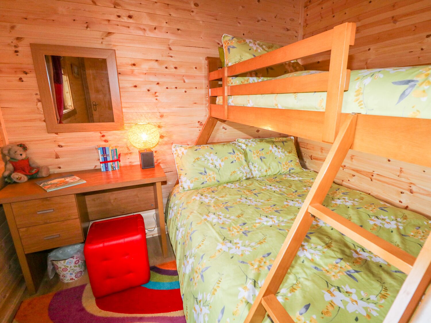 Una habitación tipo cabaña de madera con literas, un escritorio, una silla y un ventanal pequeño.