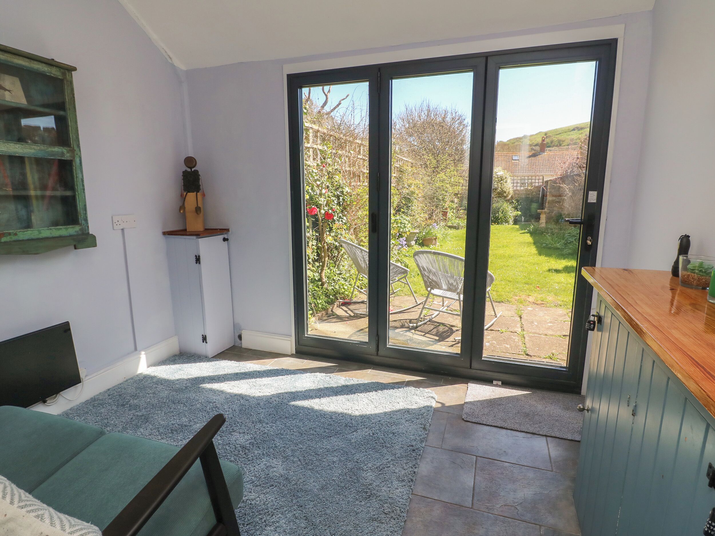 Ein Zimmer mit einer Schiebetür zu einem Garten, eine kleine Sitzecke mit einem Sessel, einem Holzschrank und einem grünen Sofa.