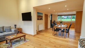 A living room with a sofa, coffee table, and TV. There's a dining area with a table and chairs. The room has wooden flooring and a view of the outdoors through a window. - Croft Cottage (Lancaster)