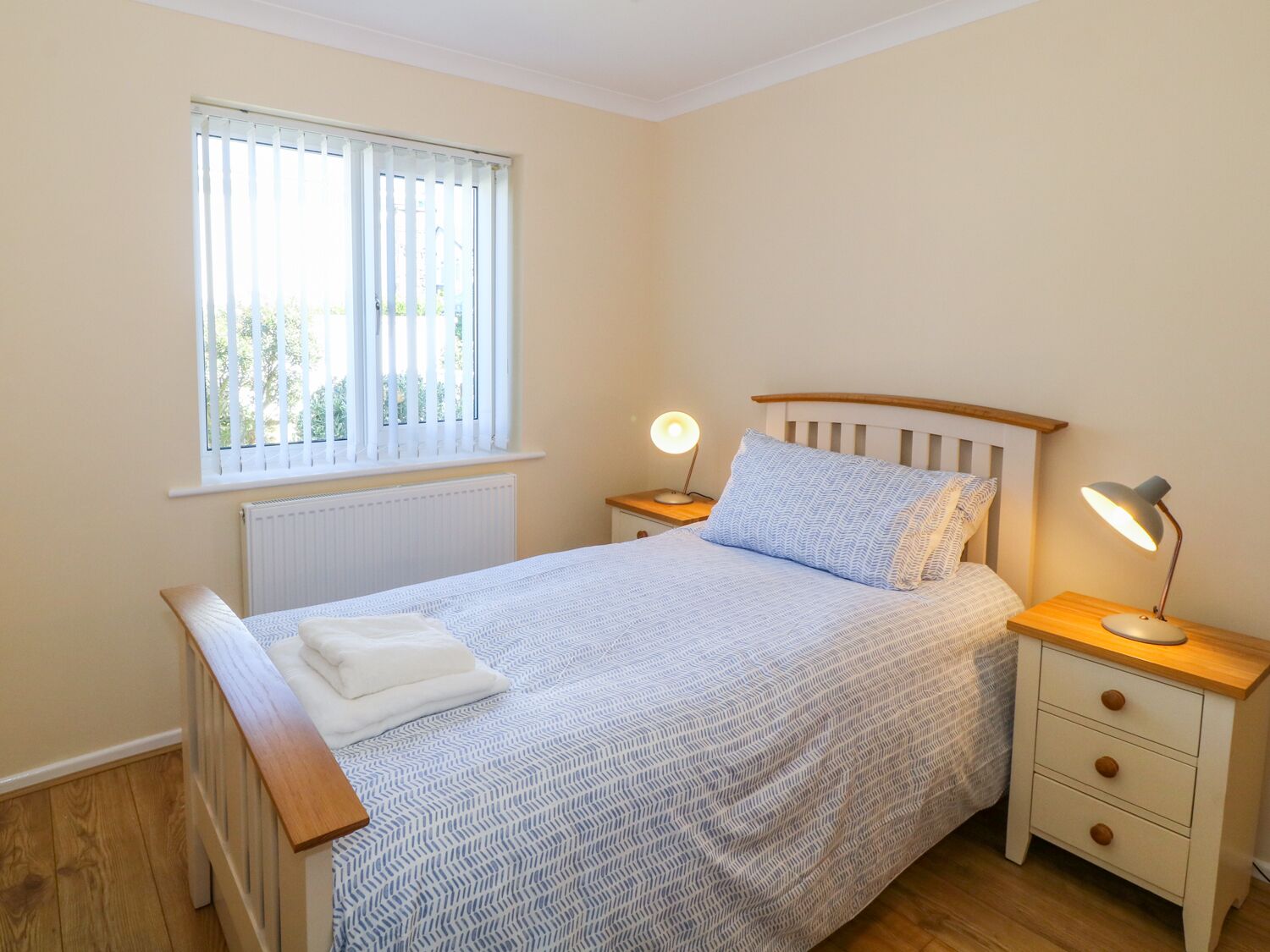 A bedroom with a bed, a window with blinds, a bedside table with a lamp, and a radiator.