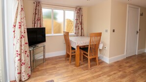 A room with a TV on a glass cabinet, a dining table with chairs, wooden flooring, and a window with curtains. - Eastdown Annexe (Totnes)