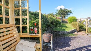 Eine Holzbank mit einer gestrickten Decke, einer Weinflasche und einem Glas auf einem Holztisch in einem Garten.