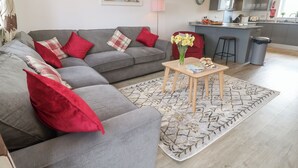 A modern living room with a grey sofa, red cushions, a small wooden table with a vase of flowers, and a patterned rug. - Sea Dreams (Marazion)
