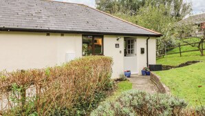 En envåningsbyggnad med ett tegeltak och en vit fasad. Det finns en dörr med ett fönster ovanför, och en liten skylt ovanför dörren med texten "12 Hand Cottage". Byggnaden är omgiven av en välskött trädgård med olika buskar och en stenlagd gångväg som leder till entrén.