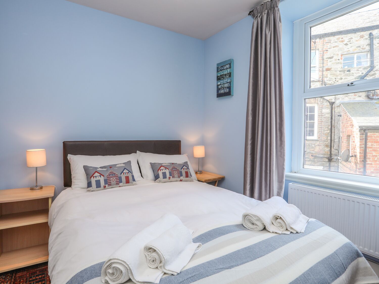 A neatly made bed with striped bedding, two bedside lamps, and a view of a brick building through a window.