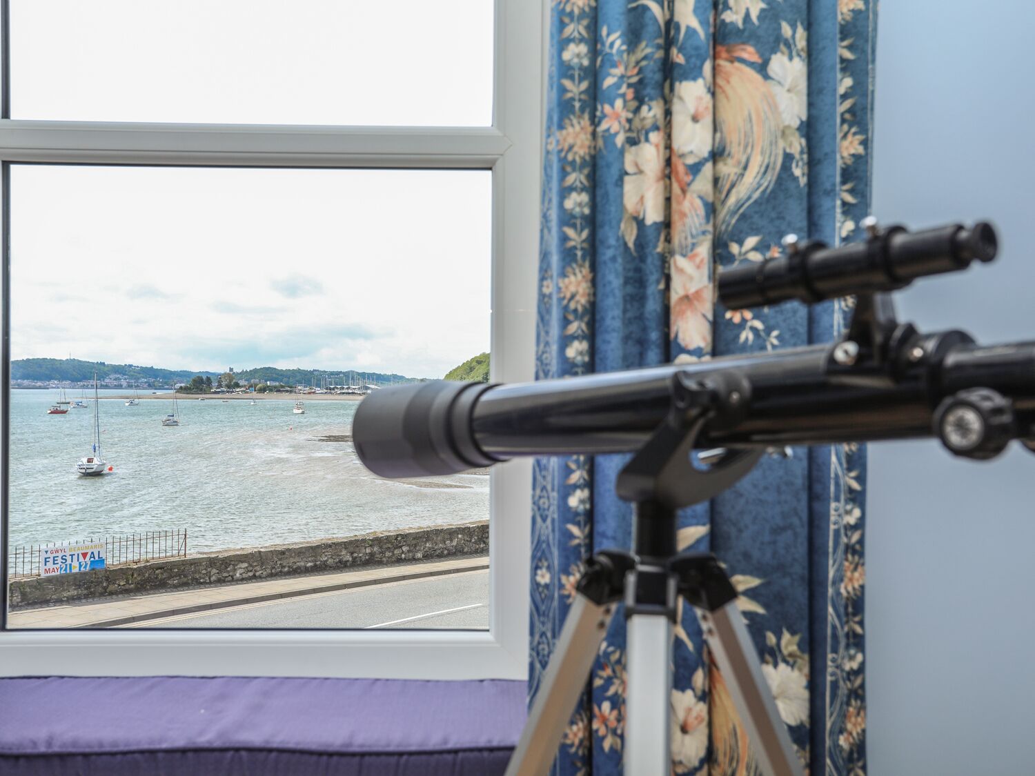 A telescope set up by a window overlooking a harbor with boats and a festival sign.