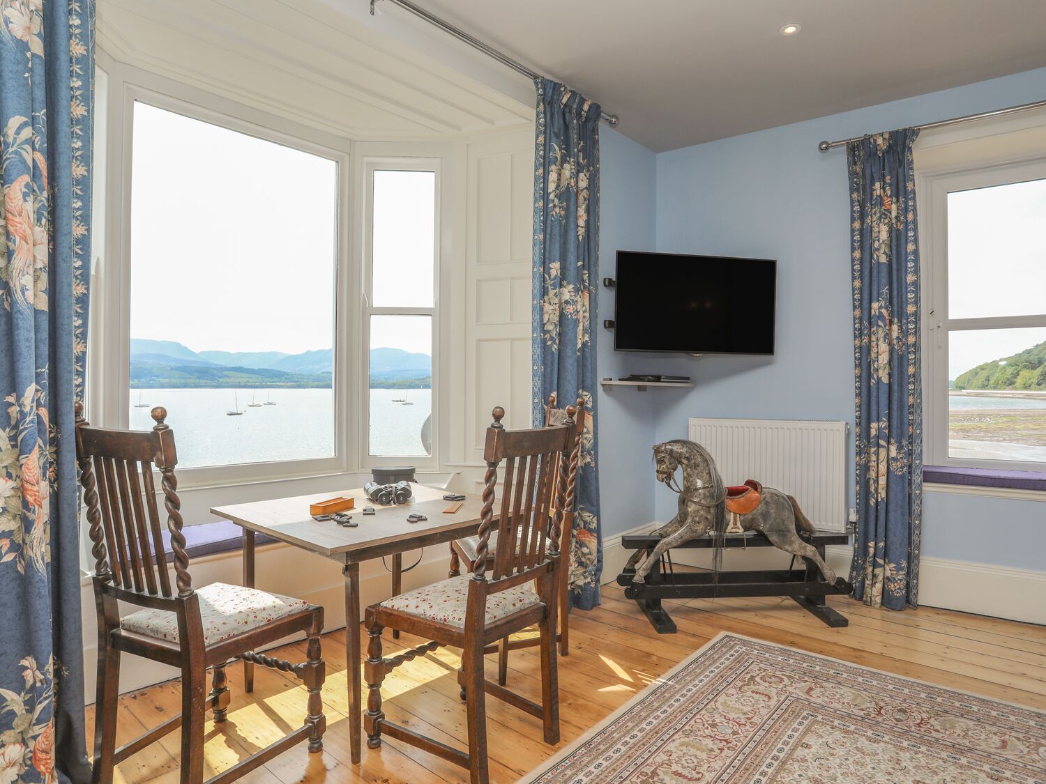 A room with a table and chairs, a television, a rocking horse, and a view of the sea.
