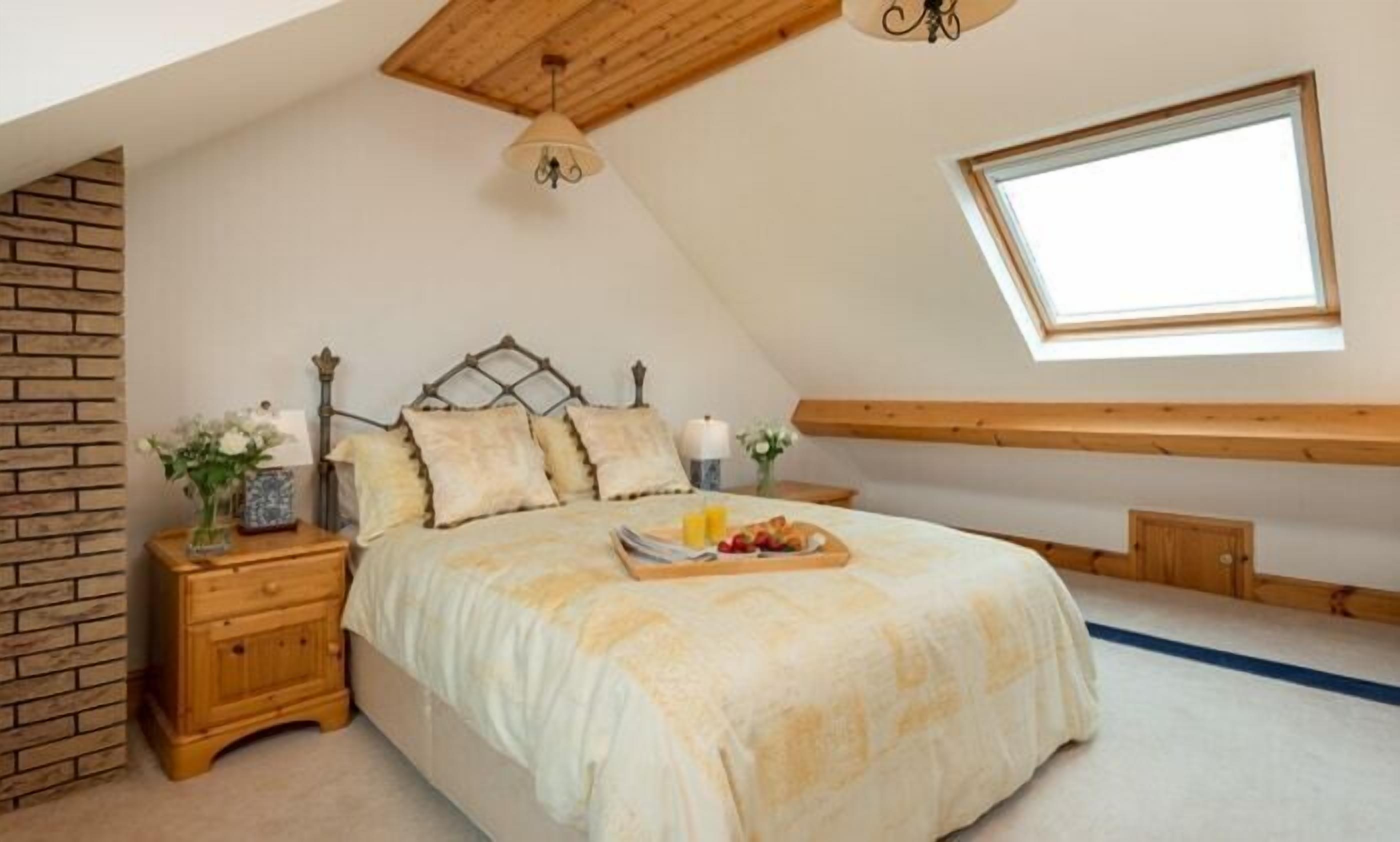 Quarto com teto inclinado, cama com cabeceira de madeira, mesa de cabeceira com luminária e janela que permite entrada de luz natural.