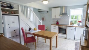 Une cuisine avec des meubles blancs, une table en bois et une chaise rouge. On y trouve un réfrigérateur, une bouilloire et une fenêtre donnant sur un paysage verdoyant.