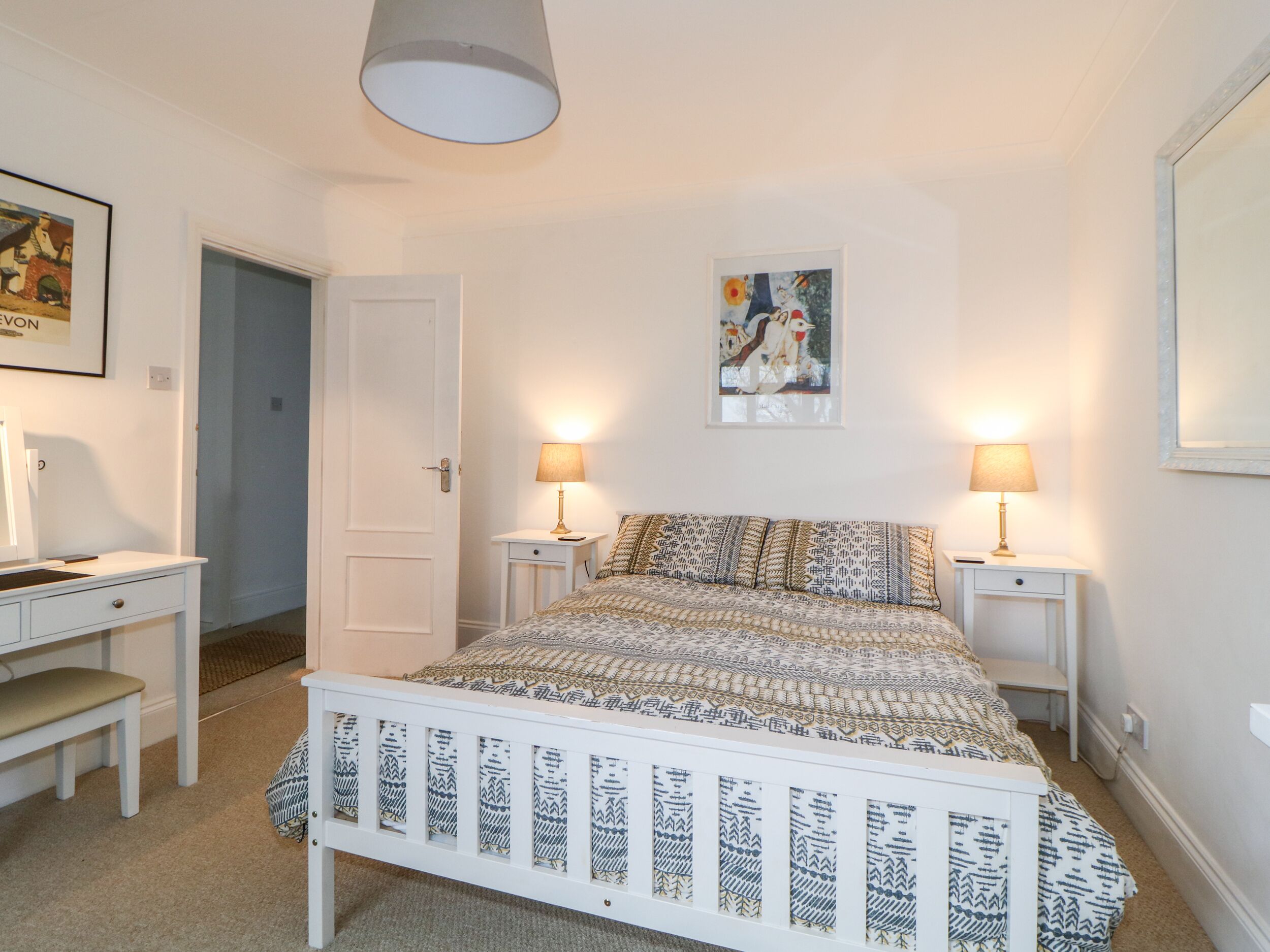 A bedroom with a white bed, two bedside tables with lamps, a desk with a chair, and a door.