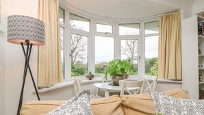 A cozy room with a curved bay window, a dining table, chairs, a floor lamp, and a bookshelf. - Hilltop (Totnes)