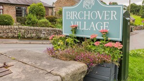 A sign for Birchover Village with a stone wall and flowering plants in front. - Andlestone (Matlock)