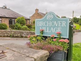 Señal de Birchover Village con un muro de piedra y plantas con flores al frente.