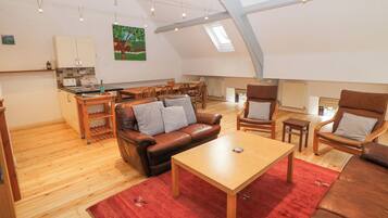 A spacious loft apartment with a high ceiling, wooden flooring, a brown leather sofa, a wooden dining table, and a kitchen area with a view of the outdoors.