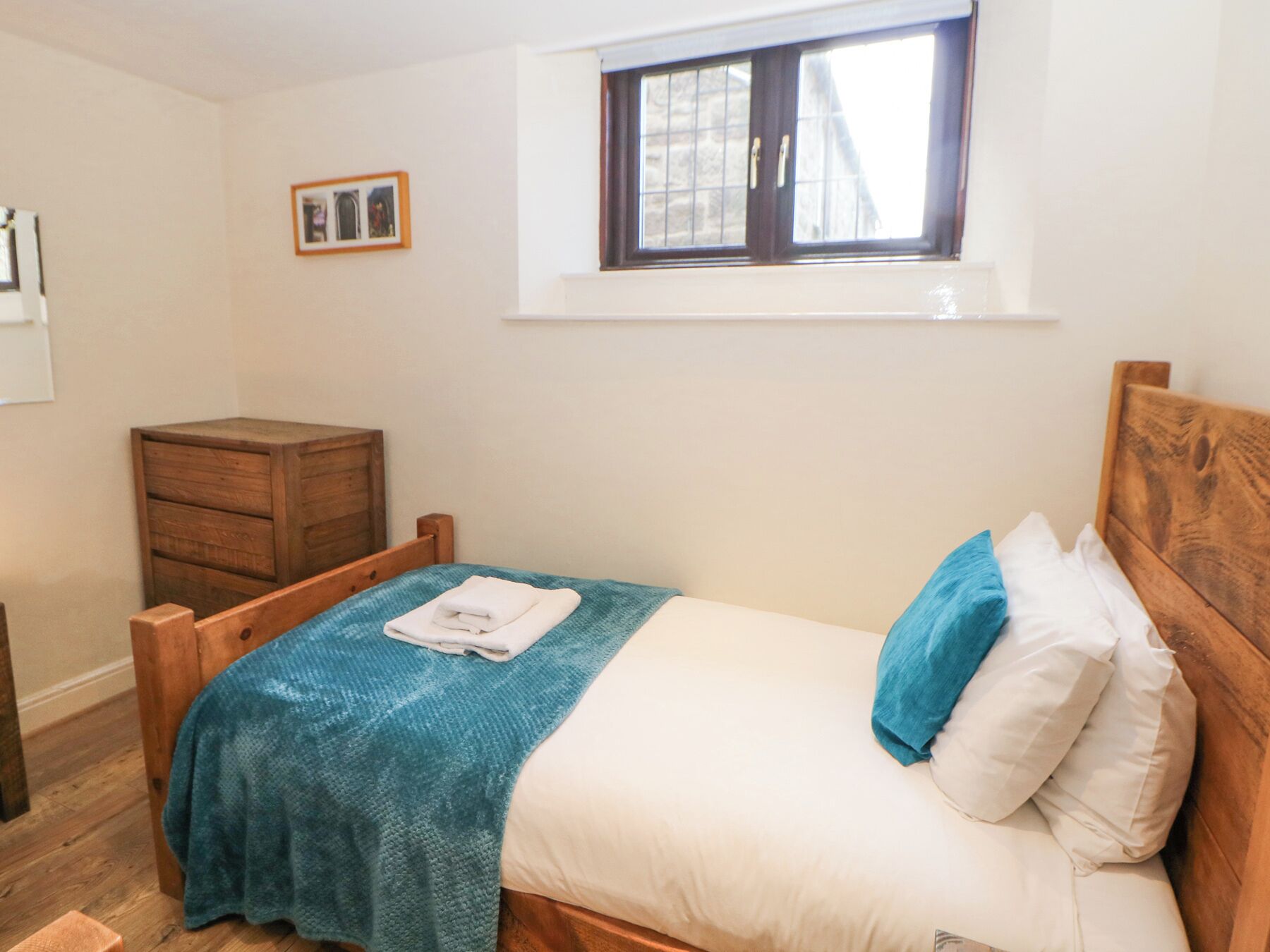 Un dormitorio con cama de madera, un pequeño aparador de madera, una ventana con cortinas e imagen enmarcada en la pared.