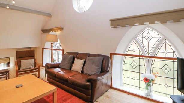 A cozy living room with a leather sofa, a wooden table, and a red rug. There is a window with a stained glass design and a round skylight. - Andlestone (Matlock)