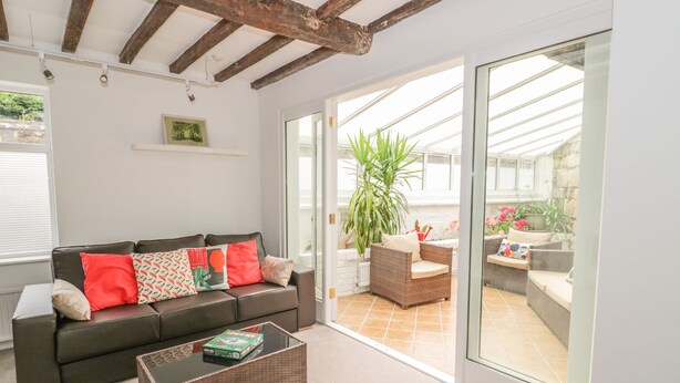 Una sala de estar acogedora con un sofá marrón oscuro, una mesa de centro de vidrio y un amplio ventanal corredero que da a un patio exterior con muebles de mimbre y plantas en macetas.