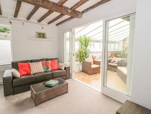A cozy living room with a dark brown sofa, a glass coffee table, and a large sliding glass door leading to an outdoor patio area with wicker furniture and potted plants. - Castle View (Warwick)